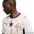 A side profile of a Black man wearing a white Ghana national team soccer jersey. The jersey features a colorful geometric pattern, a large black star in the center, and the Ghana Football Association crest. He is wearing a silver chain and looking downwar