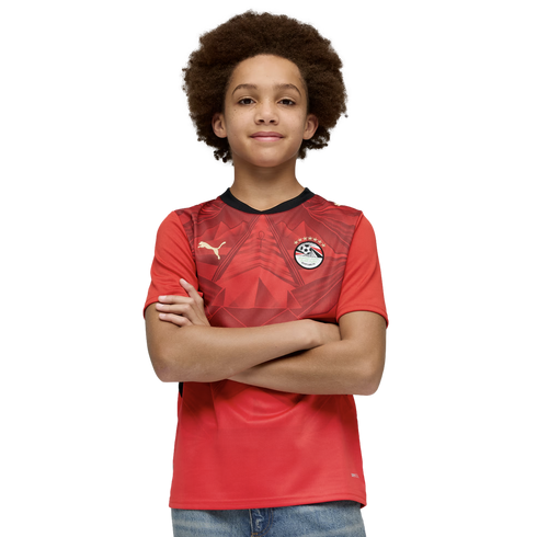 A young person with a large afro and a slight smile poses with their arms crossed. They are wearing a red Egyptian national team soccer jersey and blue jeans against a solid black background.