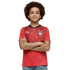 A young person with a large afro and a slight smile poses with their arms crossed. They are wearing a red Egyptian national team soccer jersey and blue jeans against a solid black background.
