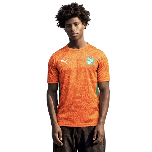 A medium shot of a young man with dark, curly hair wearing an orange Puma soccer jersey for the Côte d'Ivoire national team. The jersey features an all-over tonal leopard-print pattern, a green and white national crest on the left chest, and a white Puma