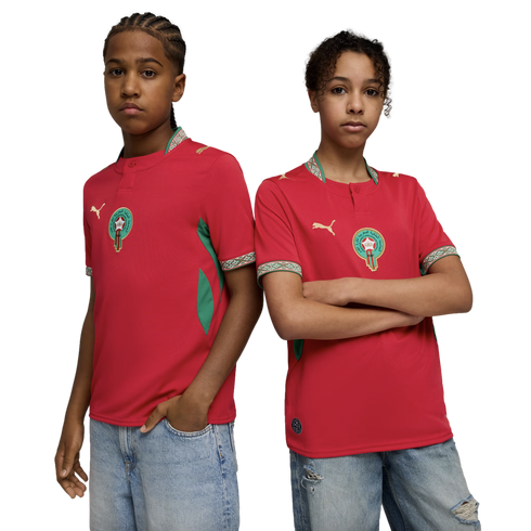 Two young people pose side-by-side against a black background, wearing red Moroccan national football team jerseys and light-wash denim jeans.