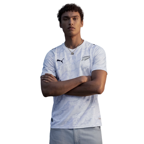 A waist-up portrait of a young man with curly hair posing with his arms crossed against a solid black background. He is wearing a white Puma jersey with a subtle, light-grey tonal swirl pattern. The jersey features a black Puma logo on the right chest and