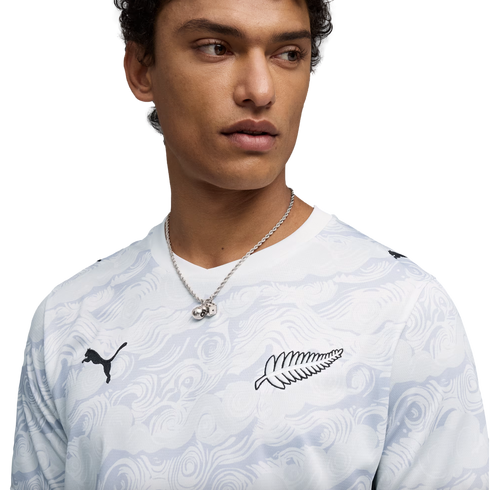 A close-up of a young man wearing a white, wavy-patterned long-sleeve Puma shirt. The shirt features a black Puma logo on the right side and a black silver fern logo on the left. He is also wearing a silver chain necklace with a dice pendant against a sol
