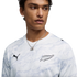 A close-up of a young man wearing a white, wavy-patterned long-sleeve Puma shirt. The shirt features a black Puma logo on the right side and a black silver fern logo on the left. He is also wearing a silver chain necklace with a dice pendant against a sol