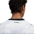 Close-up from behind of a person with dark, curly hair wearing a white jersey with a grey swirl pattern and a black collar. The word "AOTEAROA" is printed in black on the back of the jersey, just below the collar. A thin silver chain necklace is visible a