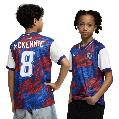 A boy and a girl model a red, blue, and white abstract-patterned soccer jersey against a black background. The boy on the left shows the back of the shirt, which features the name "MCKENNIE" and the number "8" in white. The girl on the right shows the fro