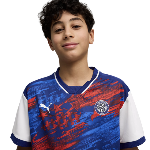 A young boy with dark curly hair and a slight smile, wearing a blue and red patterned Puma Football Club soccer jersey against a solid black background.