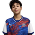 A young boy with dark curly hair and a slight smile, wearing a blue and red patterned Puma Football Club soccer jersey against a solid black background.