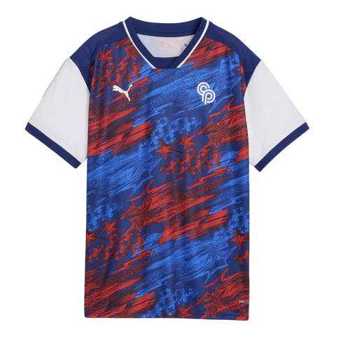 A short-sleeved Puma soccer jersey with a patriotic red, white, and blue theme. The main body of the shirt features an energetic, abstract pattern of blue and red brushstrokes and stars on a navy background. The jersey has a navy blue V-neck collar with w