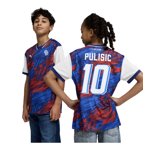 A studio portrait against a black background shows two young children modeling a blue and red Puma soccer jersey. The boy on the left faces forward, showing the front of the jersey with its abstract star pattern and Puma logo. The girl on the right faces
