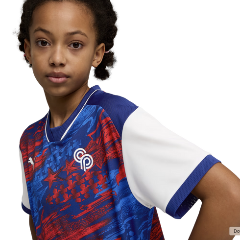 A young girl with brown skin and dark curly hair looks directly at the camera against a solid black background. She wears an athletic jersey with a blue, red, and white graphic pattern featuring stars, a white "CP" logo on the left chest, and a solid whit