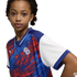 A young girl with brown skin and dark curly hair looks directly at the camera against a solid black background. She wears an athletic jersey with a blue, red, and white graphic pattern featuring stars, a white "CP" logo on the left chest, and a solid whit