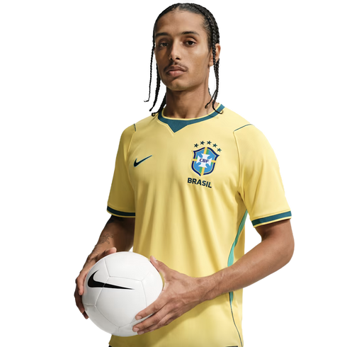 A man with braided hair wearing a yellow Nike Brazil national football team jersey, holding a white soccer ball against a solid black background.
