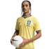 A man with braided hair wearing a yellow Nike Brazil national football team jersey, holding a white soccer ball against a solid black background.