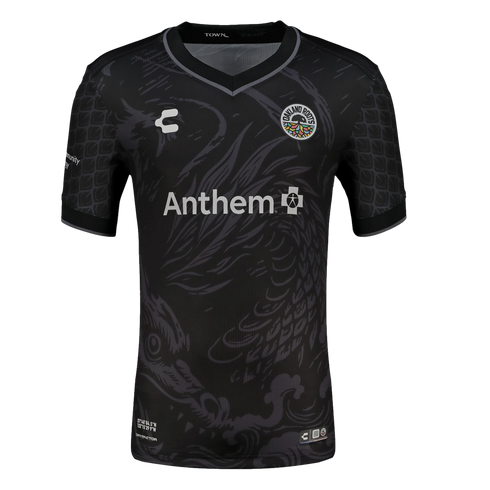A black short-sleeved soccer jersey featuring a large, subtle grey dragon illustration across the front. The jersey includes the colorful Oakland Roots SC crest on the chest, a white Anthem sponsor logo in the center, and a repeating grey scale pattern on