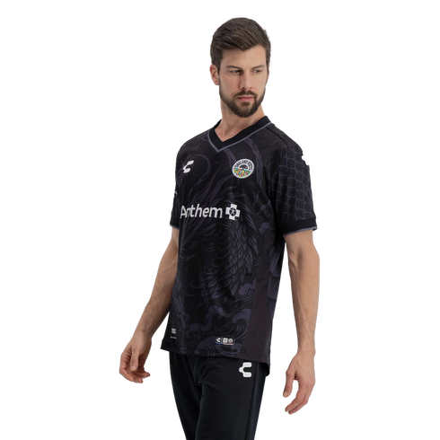 A man models an Oakland Roots SC soccer jersey against a solid black background. The black V-neck jersey features tonal grey wavy patterns on the front, textured scale-like patterns on the sleeves, the team's circular crest on the chest, and an Anthem spo