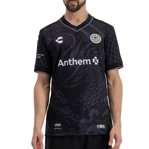 A man wearing a black Oakland Roots soccer jersey featuring a subtle tonal dragon graphic. The jersey has a white "Anthem" sponsor logo across the center, the team crest on the left chest, and geographic coordinates printed at the bottom left.