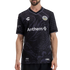 A man wearing a black Oakland Roots soccer jersey featuring a subtle tonal dragon graphic. The jersey has a white "Anthem" sponsor logo across the center, the team crest on the left chest, and geographic coordinates printed at the bottom left.