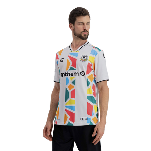 A man models a white Oakland Roots soccer jersey featuring a vibrant, multi-colored geometric mosaic pattern in shades of red, yellow, orange, blue, and green. The word "Anthem" is printed in black across the chest, and the jersey includes a black-and-whi