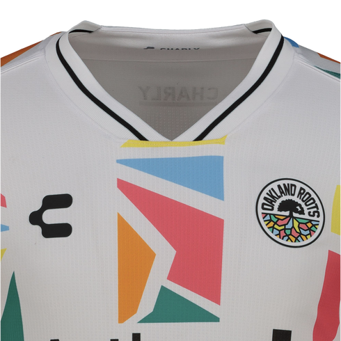 Close-up of a white Oakland Roots SC soccer jersey by Charly, featuring a large, colorful geometric pattern on the chest and the team's circular tree crest.