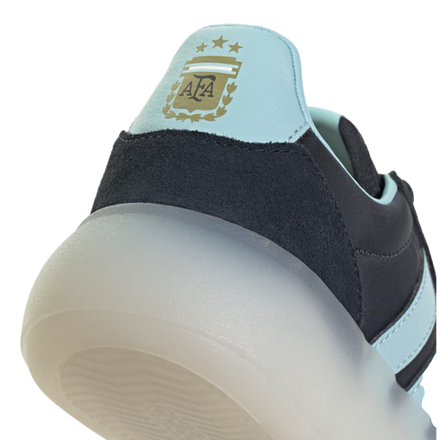 A close-up shot focusing on the heel of an Adidas shoe, featuring a light blue panel with the gold Argentina Football Association (AFA) crest and three stars. Below the blue panel is a dark navy textured material and a thick, semi-translucent white sole,