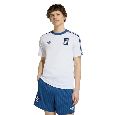 A man models a white Adidas t-shirt and blue striped shorts, both with the Greek national team crest, against a black background.