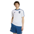 A man models a white Adidas t-shirt and blue striped shorts, both with the Greek national team crest, against a black background.