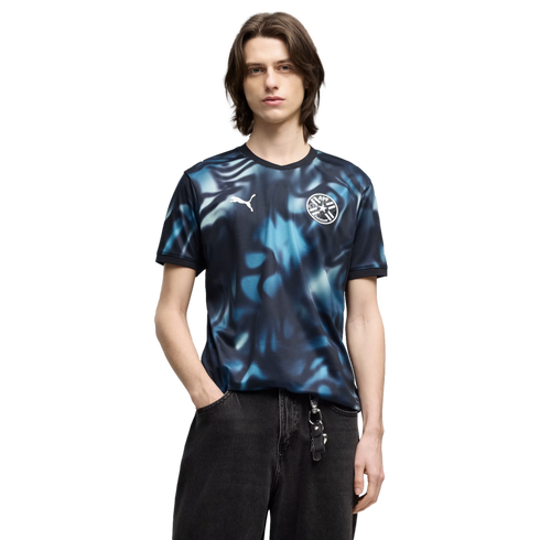 A man with brown hair wears a blue and black abstract Puma jersey with the Paraguay football crest and black jeans against a solid black background.