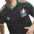 Close-up of a person wearing a black Mexico football jersey with green, white, and red accents, the national team crest, and a colorful Adidas trefoil logo.