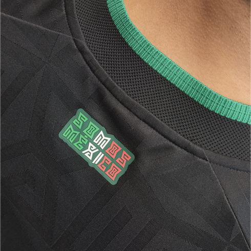 Close-up of a black jersey with a green-ribbed collar and a "SOMOS MÉXICO" patch in green, white, and red. The fabric has a subtle geometric pattern.