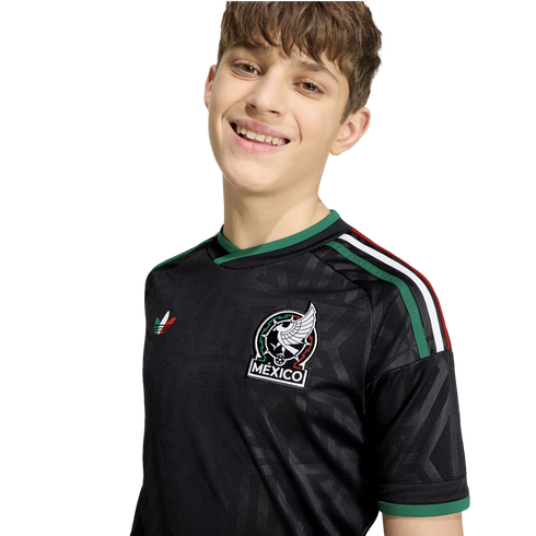 A smiling young person wearing a black Mexico national football team jersey with green accents and the official team crest, set against a plain white background.