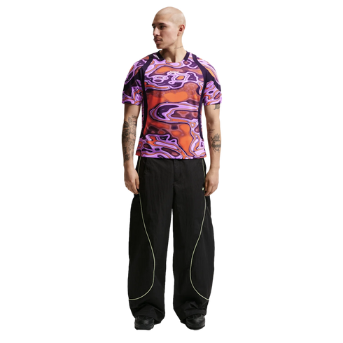 A man wearing a purple and orange psychedelic-patterned t-shirt and black wide-leg pants with neon piping, standing against a white background.