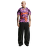 A man wearing a purple and orange psychedelic-patterned t-shirt and black wide-leg pants with neon piping, standing against a white background.