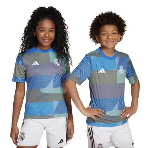 Two children smile while posing in matching blue and teal geometric-patterned Real Madrid soccer jerseys and white shorts.