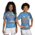 Two children smile while posing in matching blue and teal geometric-patterned Real Madrid soccer jerseys and white shorts.