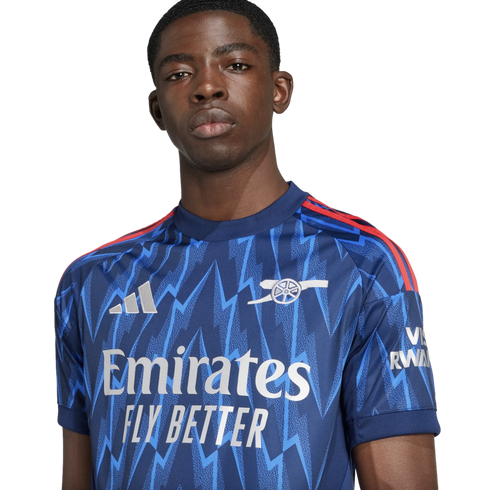 Portrait of a young Black man wearing a blue Arsenal football jersey with a zigzag pattern, posed against a black background.