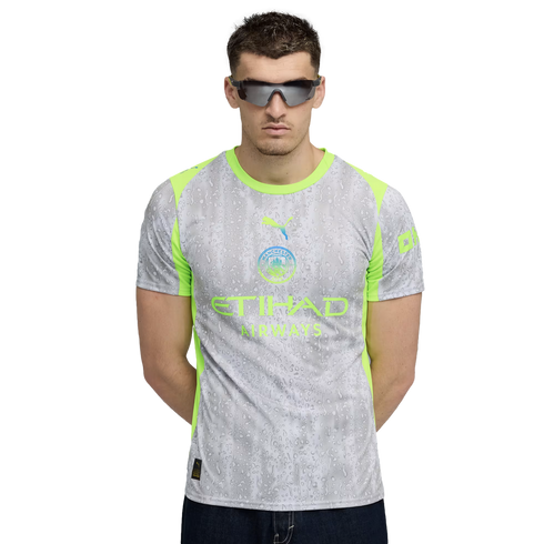 A man in dark sunglasses stands against a black background wearing a grey and lime green Manchester City jersey with a water droplet pattern.