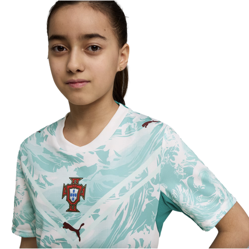A young girl with dark hair tied back wears a white and teal patterned Portugal national football team jersey with the FPF crest and dark red Puma logos. She looks directly at the camera against a solid black background.