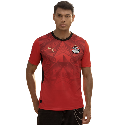 A man wearing a red Egypt national football team home jersey by Puma, featuring subtle geometric patterns and the team crest, standing against a plain black background.