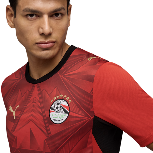 A close-up portrait of a man wearing a red Egypt national football team jersey by Puma. The jersey features an intricate geometric pattern, the Egyptian FA crest with seven gold stars, and gold Puma logos. He looks slightly off-camera against a solid blac