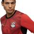 A close-up portrait of a man wearing a red Egypt national football team jersey by Puma. The jersey features an intricate geometric pattern, the Egyptian FA crest with seven gold stars, and gold Puma logos. He looks slightly off-camera against a solid blac