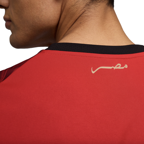 A close-up photograph shows the upper back and neck of a person wearing a red sports jersey. The jersey features a black collar and a stylized gold inscription of the word "Egypt" in Arabic (مصر) on the upper back.