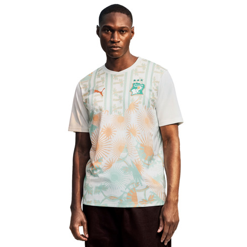 A Black man stands against a plain black background, wearing a white Côte d’Ivoire national football team jersey by Puma. The jersey features an orange Puma logo on the right chest and the team crest on the left, with a light green and orange graphic patt