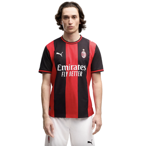 A man with long dark hair wearing a red and black striped AC Milan soccer jersey and white shorts, standing against a plain white background.