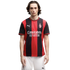 A man with long dark hair wearing a red and black striped AC Milan soccer jersey and white shorts, standing against a plain white background.