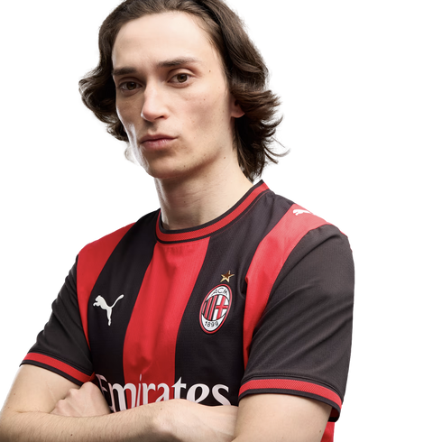 A man with shoulder-length brown hair and arms crossed, wearing a red and black AC Milan soccer jersey with a white Puma logo against a plain white background.