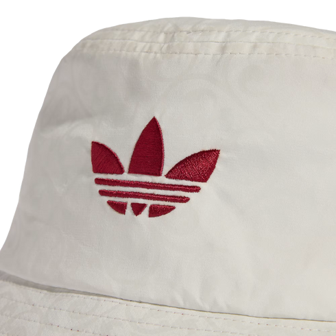 A close-up of a white bucket hat featuring a red embroidered Adidas trefoil logo on the front crown. The fabric has a subtle, repeating tonal pattern weave.