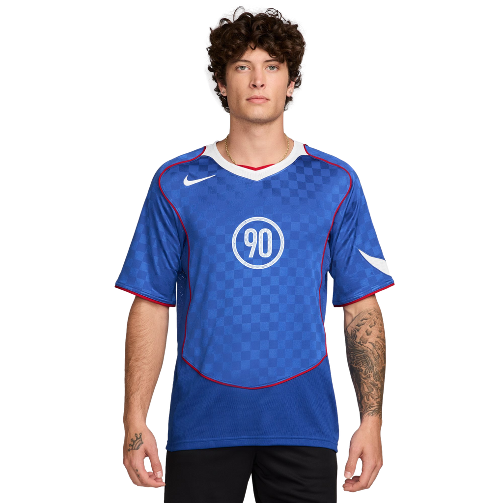A man with curly hair wearing a blue Nike Total 90 soccer jersey with a white "90" logo and checkered pattern.