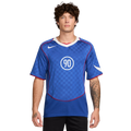 A man with curly hair wearing a blue Nike Total 90 soccer jersey with a white "90" logo and checkered pattern.