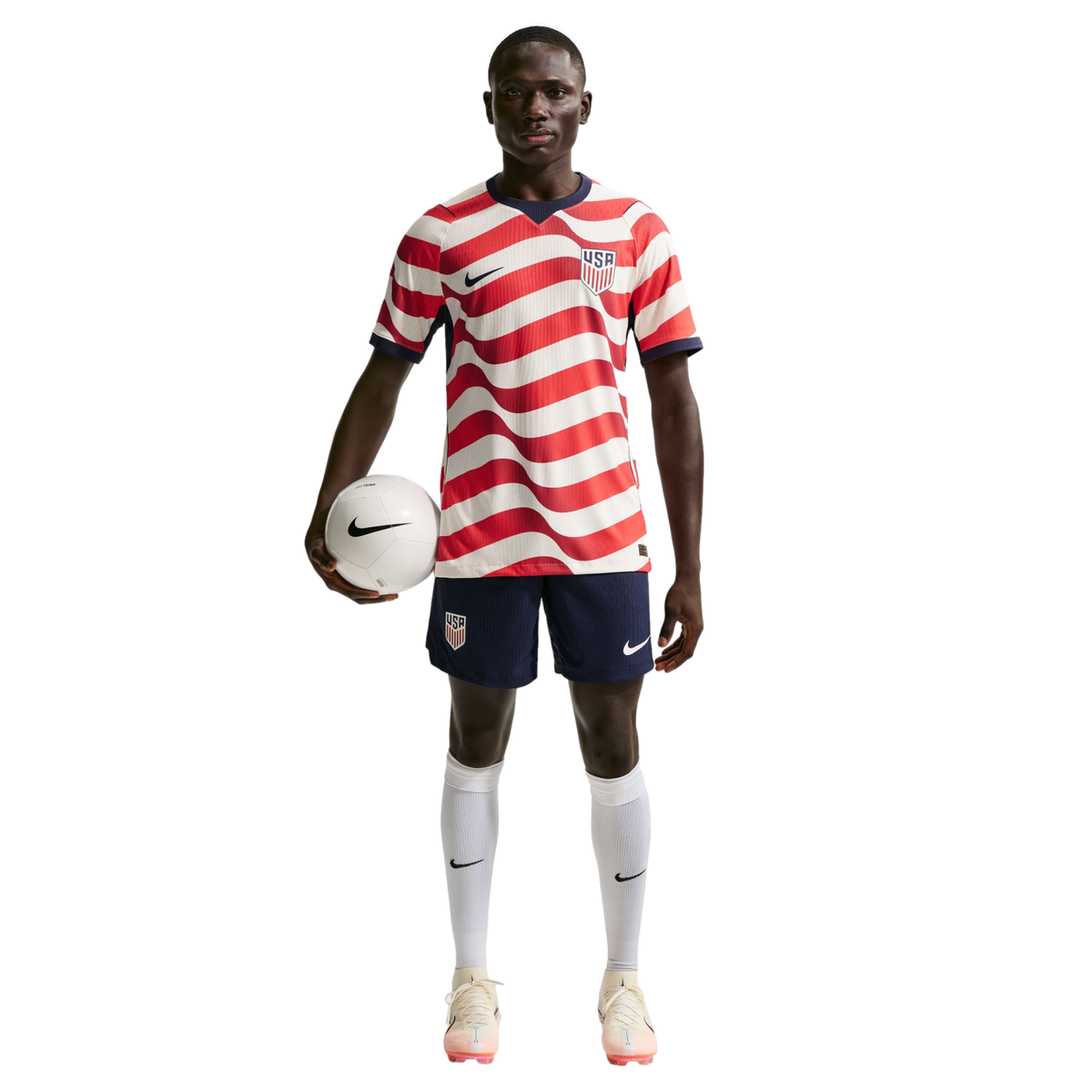 A male soccer player in a wavy red and white USA jersey and navy shorts, holding a soccer ball under his arm against a black background.